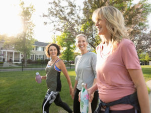 Three middle-aged women walking through a neighborhood park, smiling, talking, wearing workout gear and holding water bottles.