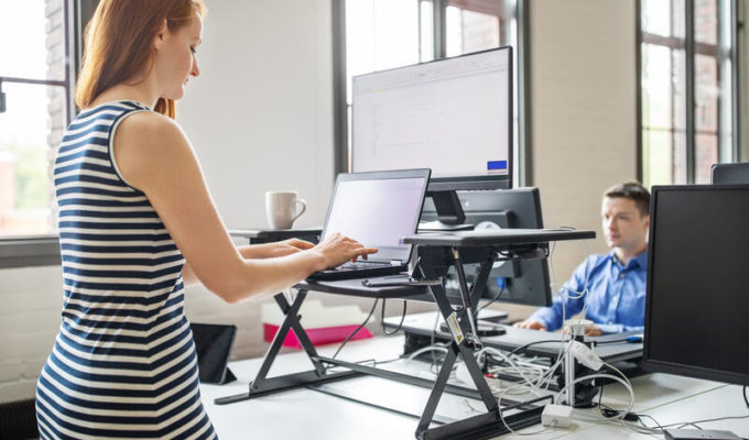 Female professional working at her standing desk with male colleague seated working at the back.
