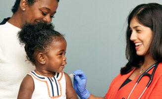 Toddler girl on mom's lap smiles at medical provider who is preparing her arm for a routine childhood immunization.