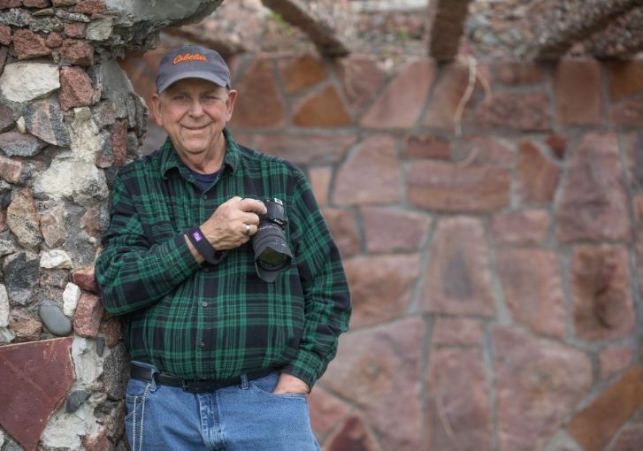 Photographer Chuck Taylor stands with his camera in a pergola-style shelter made of pink stone.