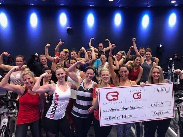 A group of women in workout gear flexes while holding a big check for $615 made out to the American Heart Association.