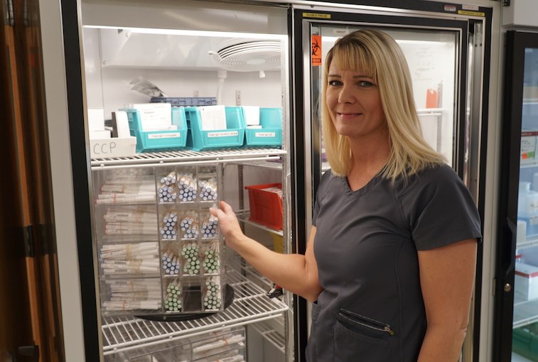 A lab employee stands next to an open refrigerator with test tubes organized on shelves.
