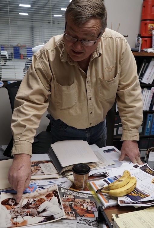 An older man with glasses stands over a desk covered with papers. He points to a photo in an open magazine.