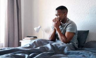 A handsome young man sitting in his bed and blowing his nose while suffering from the flu.