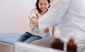 Cheerful girl sitting in an urgent care exam room and holding up her arm while being examined by a doctor.
