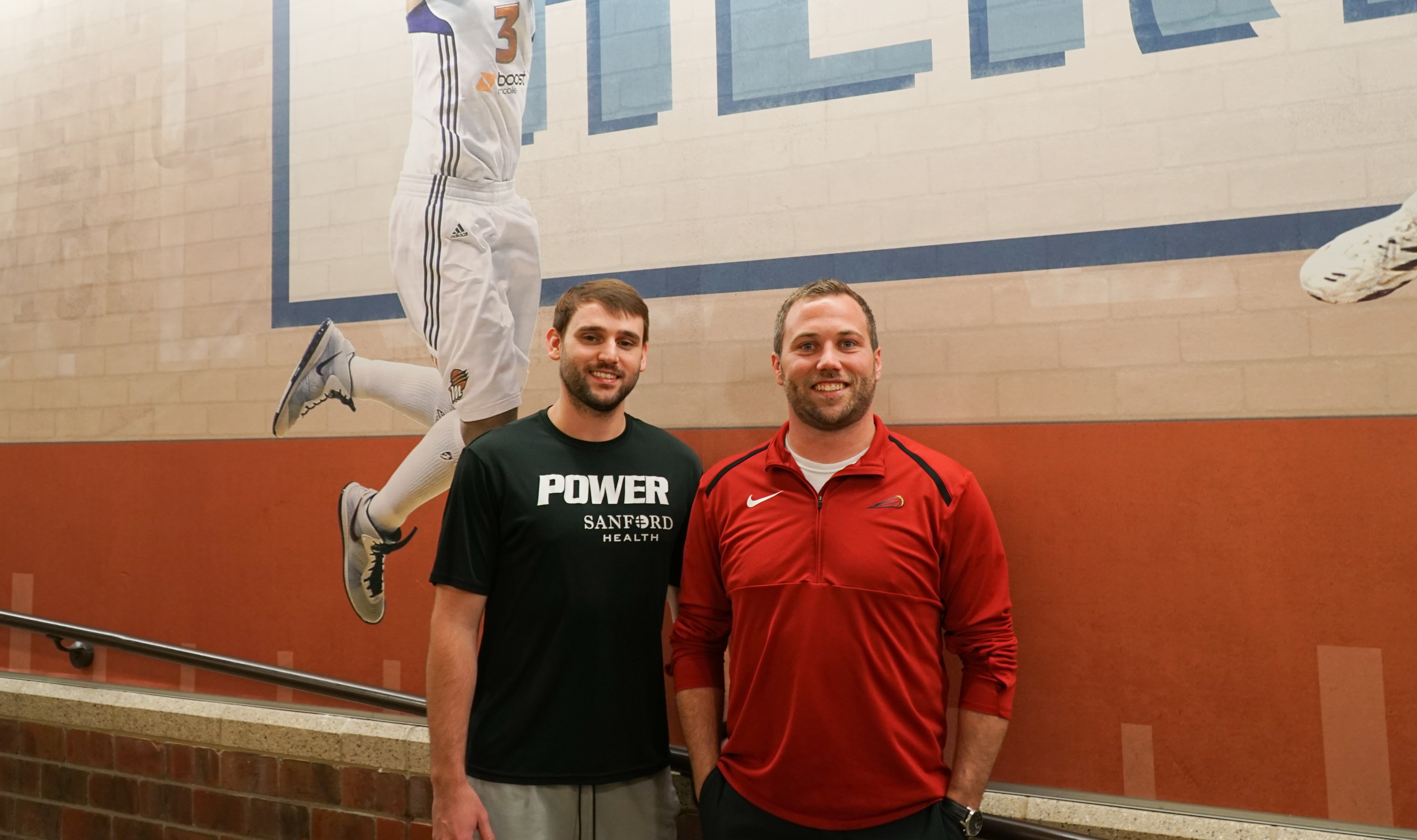 Two Sanford Health employees smile while standing against a gym wall with a basketball mural painted on it.