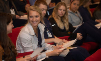 A young nurse smiles at a classmate while a row of other nurses looks on. They are sitting in an auditorium-style classroom as part of the Sanford Nurse Residency Program.