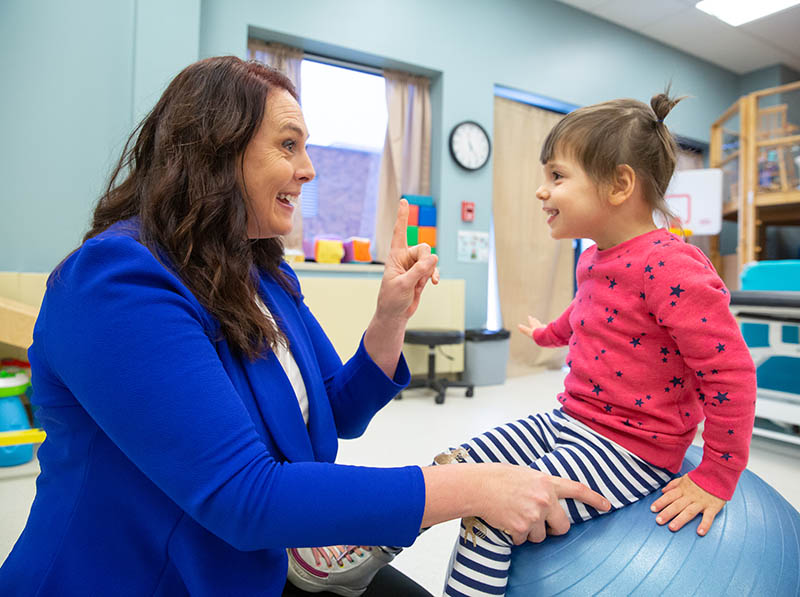 A woman smiles and holds up one finger while helping a smiling little girl balance on top of a ball at Sanford Bemidji Children's Therapy.