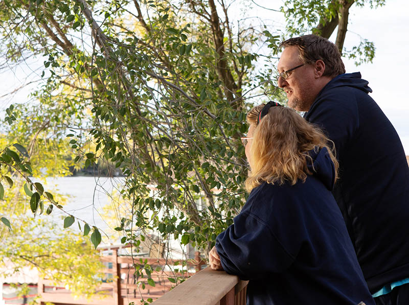 Ken and Mindy Santema stand on their deck looking through leafy green trees to a lake.