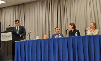 Four speakers share a stage with blue and gray curtains.