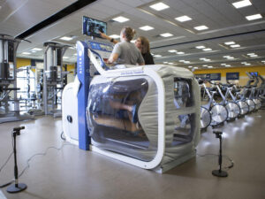Woman runs on anti-gravity treadmill which puts her lower body inside a plastic cube. A trainer stands at her side.