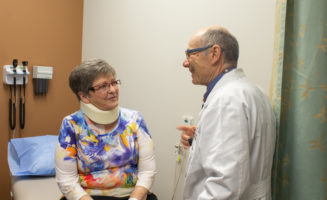 Cancer survivor Darlene Kloeppner, seated, wears a neck brace while talking with her doctor in an exam room.