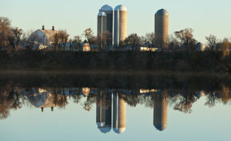 A farmstead with grain bins and a barn stands in the background, reflected in a body of water in the foreground. Flooding has made farming a challenge.