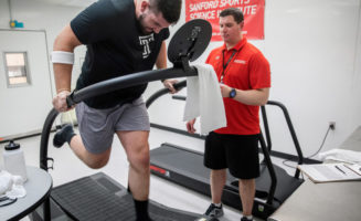 College football player and Rep 1 Sports athlete Matt Hennessy runs on a treadmill with a Sanford Health staff member timing him. A red sign in the background says Sanford Sports Science Institute.