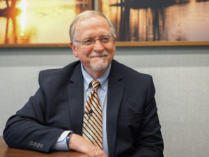 Gene Hoyme sits with his arm on a table smiling at an off-camera interviewer