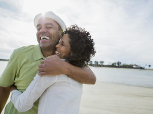 A happy, healthy mid-40s couple hugs on a sunny beach.