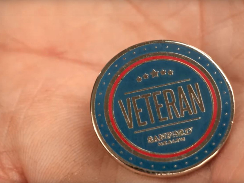 Blue, red and gold circular pin in the palm of a man's hand says VETERAN - Sanford Health.
