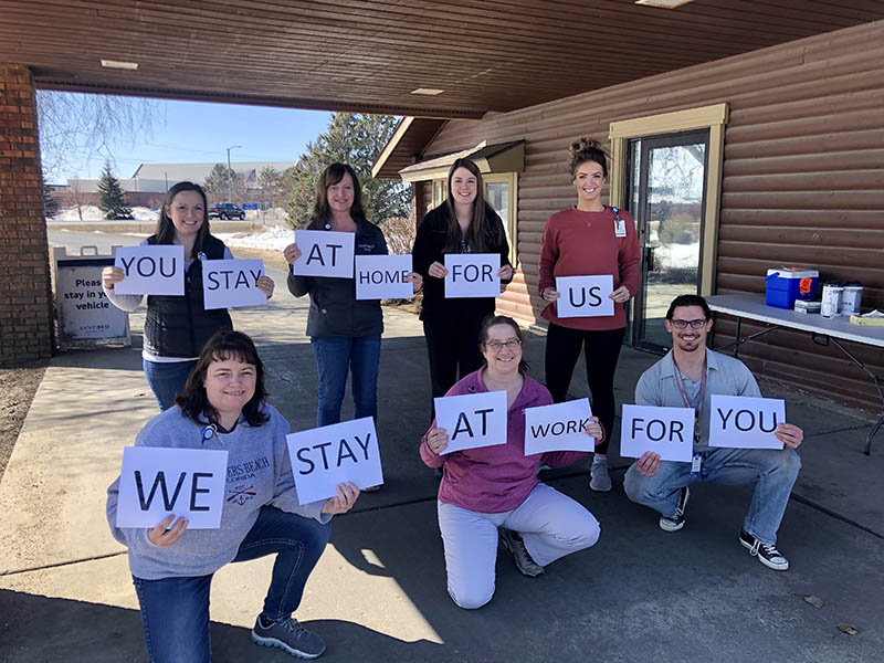 A Sanford Health COVID-19 testing team holds printed pieces of paper that say YOU STAY AT HOME FOR US, WE STAY AT WORK FOR YOU.