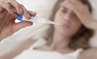 A woman with her hand on her forehand holds up a thermometer. Answers to questions about Sanford Health Plan's coverage of testing costs and more related to coronavirus.