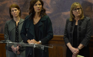 Allison Suttle, Kristi Noem and Kim Malsam-Rysdon stand listening to questions at a clear podium.