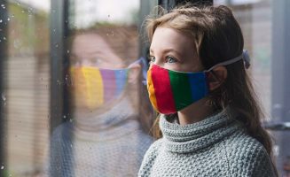A girl in a rainbow face mask looks out the window on a cloudy day.