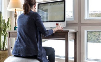 Social distancing: Woman works from home sitting on exercise ball at her desk faces a computer screen and large windows.