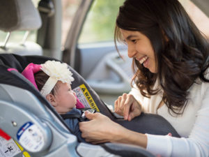 A mom smiles at her baby as she buckles her into a car seat. Experts caution against placing children into forward-facing car seats too soon.