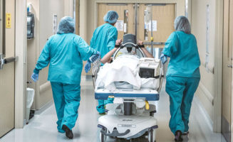 A patient appears to be wheeled down a hallway by three nurses, with a LUCAS mechanical CPR device on his chest.