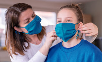 A mom wearing a blue face mask places one on her daughter's face as well.