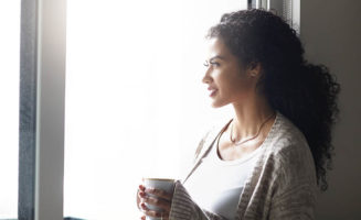 The silhouette of a woman standing in front of a window holding a cup of coffee, in a possible pose of mindfulness