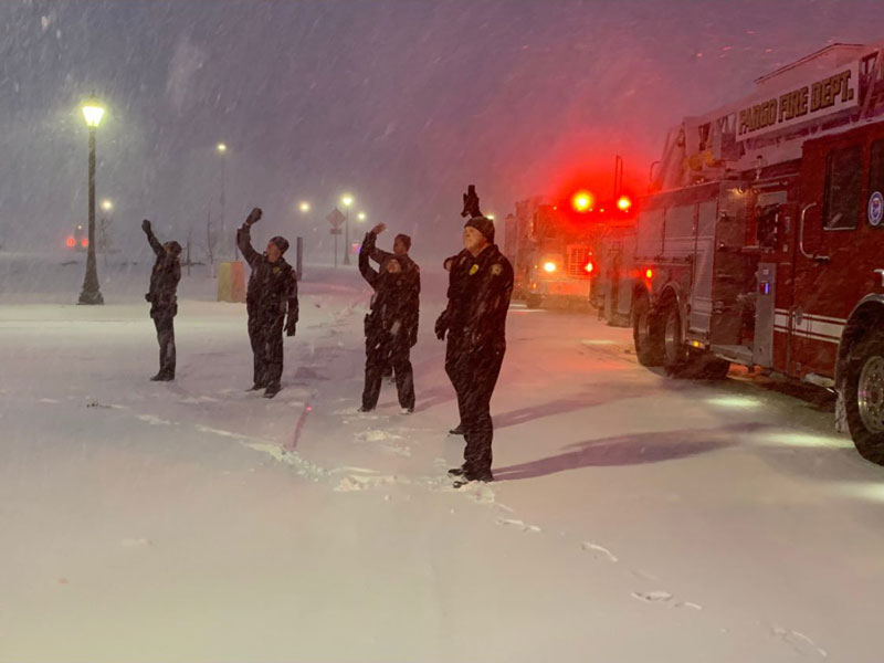 Uniformed first responders wave to hospital patients while standing in the snow. The blue and red lights of their cars are shining in the night.