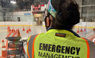 A woman in a mask and a yelow Emergency Management vest looks out at the Worthington Ice Arena set up for drive-thru COVID-19 testing.