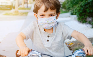 Caucasian little boy riding a bicycle wearing a protective mask.