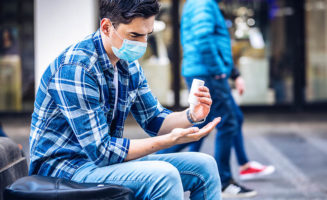 A young man sits on a bench outdoors wearing a face mask and applying sanitizer in an illustration of life's new normal
