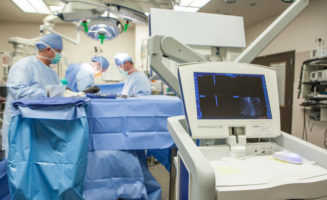 Dr Geoffrey Haft and two other surgeons in personal protective equipment work on a patient at a table draped in blue. A computer in the foreground shows an X-ray of a spine..