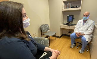 Caucasian woman in a mask talks to a caucasian fertility doctor in a mask as they sit in an exam room.