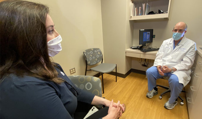 Caucasian woman in a mask talks to a caucasian fertility doctor in a mask as they sit in an exam room.