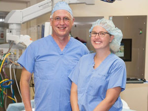 Father and daughter doctors and OB/GYNs pose in scrubs in a Sanford Health hospital room.