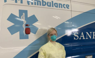 Stephanie Tegtmeier, paramedic, stands by an ambulance in a face mask and paper gown. The graphics on the side of the ambulance in blue and white say F-M AMBULANCE and SANFORD HEALTH.