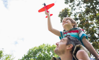 A boy sits on his dad's shoulders outside while flying a toy airplane.