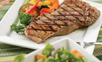 Extra large steak with roasted vegetables on the side of a white plate on a colorful tablecloth outside.