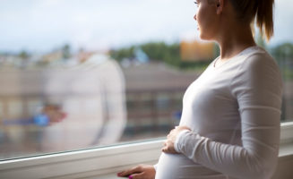 Thoughtful pregnant woman looks out the window home. Holding a hand on the tummy. Mental health and pregnancy.