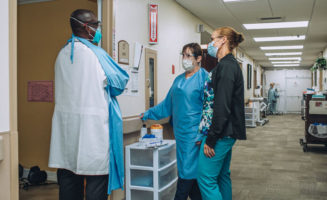 Three Good Samaritan Society employees in PPE stand talking outside a nursing home resident's room.