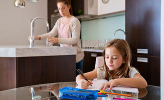 A girl works on homework at the kitchen table while her mom stands at the sink after her daughter comes home from school