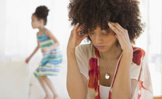 A woman holds her hands to her forehead in stress while her daughter plays in the background