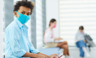 Boy wearing mask sitting in medical waiting room holding gadget looking at camera