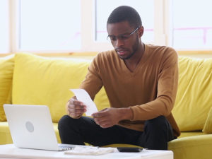 Stressed man on couch looks at a receipt while working on paying bills online.
