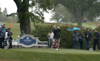 Fans stand behind rope lines as John Daly swings near a sign for the 2020 Sanford International golf tournament sign.