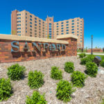 A brick sign for Sanford is nestled among landscaping outside Sanford Medical Center Fargo, towering in the background on a sunny day.