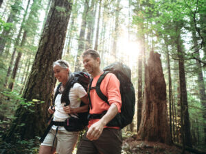 Active sporty mature couple hiking through forest with sun in background.